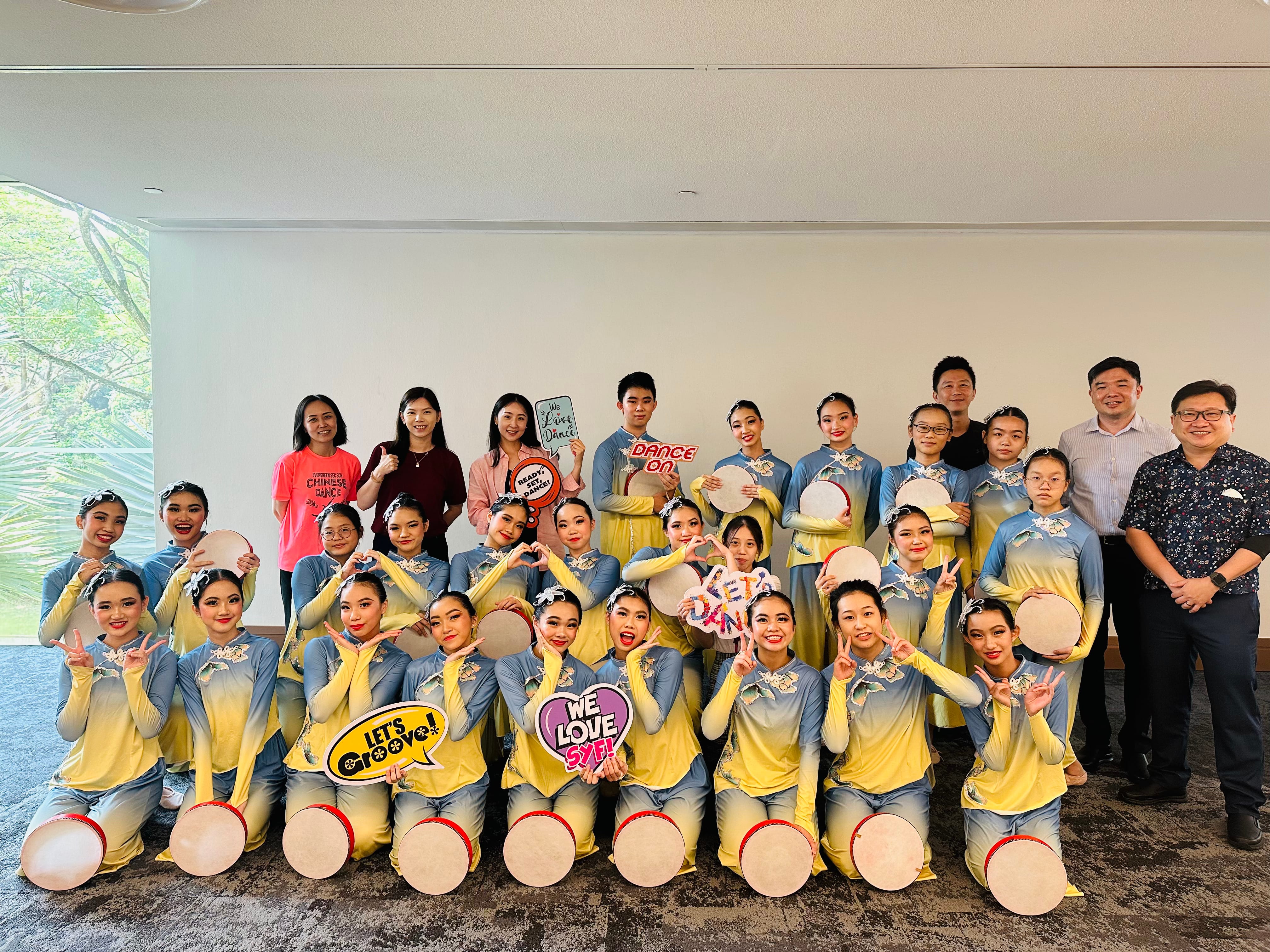 Group photo of dancers in blue-yellow outfits holding round fans, with adults standing behind them.